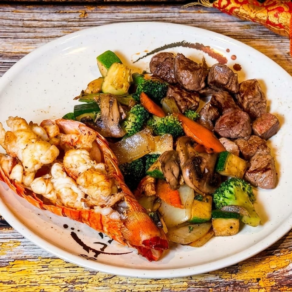 Lobster tail and steak with mixed vegetables on a white plate, next to a decorative fan.